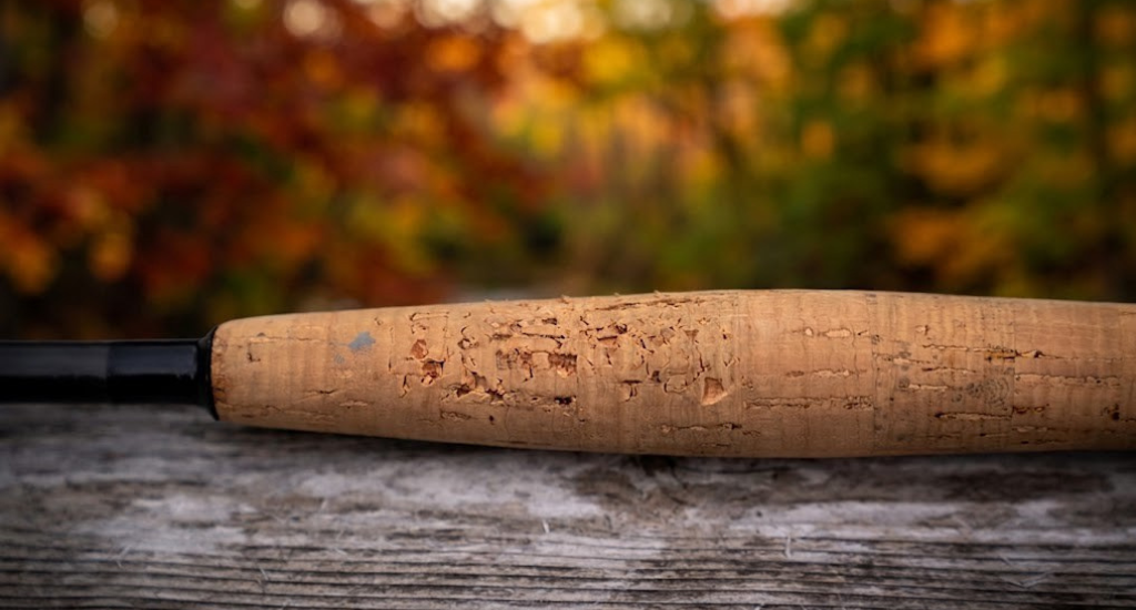 Close-up of a damaged cork handle on a fishing rod resting on a weathered wooden surface, featuring deep pits and gouges against a soft, autumnal bokeh background.