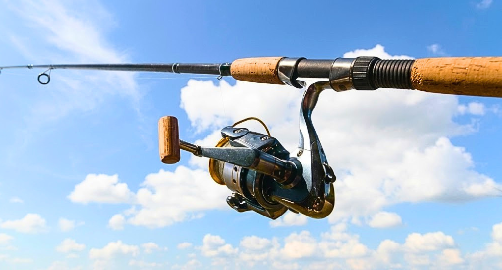 A spinning fishing rod and reel held against a bright blue sky with white clouds. The reel is silver and gold, and the rod has a classic light-colored cork handle.