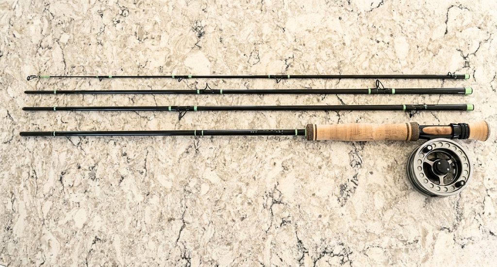 A four-piece fly fishing rod disassembled and laid out horizontally next to its reel on a textured surface.