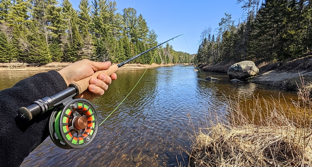 A first-person view of a hand holding a fly fishing rod and reel over a flowing river lined with pine trees.