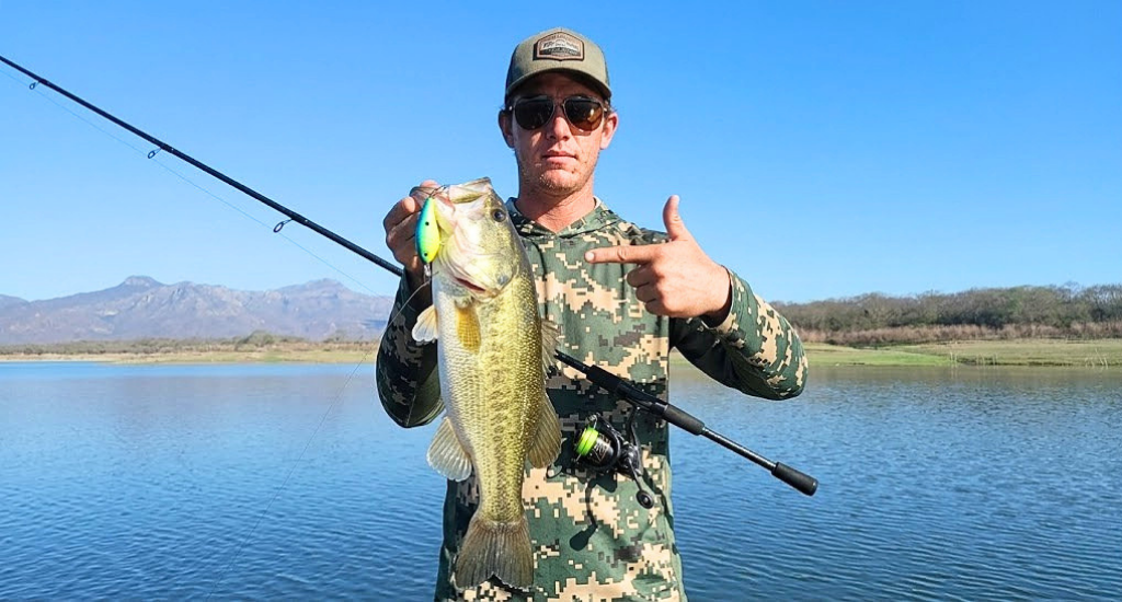 An angler holding a caught bass with a lure in its mouth in front of a lake with mountains in the background.