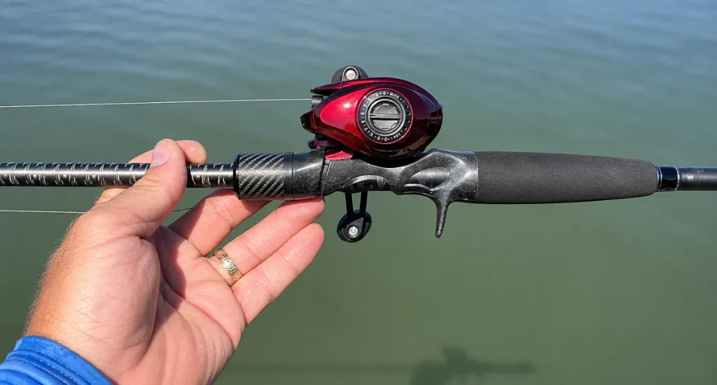 Hand holding a carbon fiber baitcasting fishing rod with a red low-profile reel over open water.