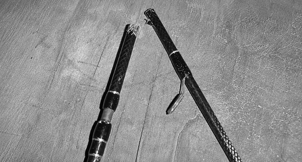Black and white top-down view of a snapped fishing rod blank lying on a wooden workbench next to a line guide.
